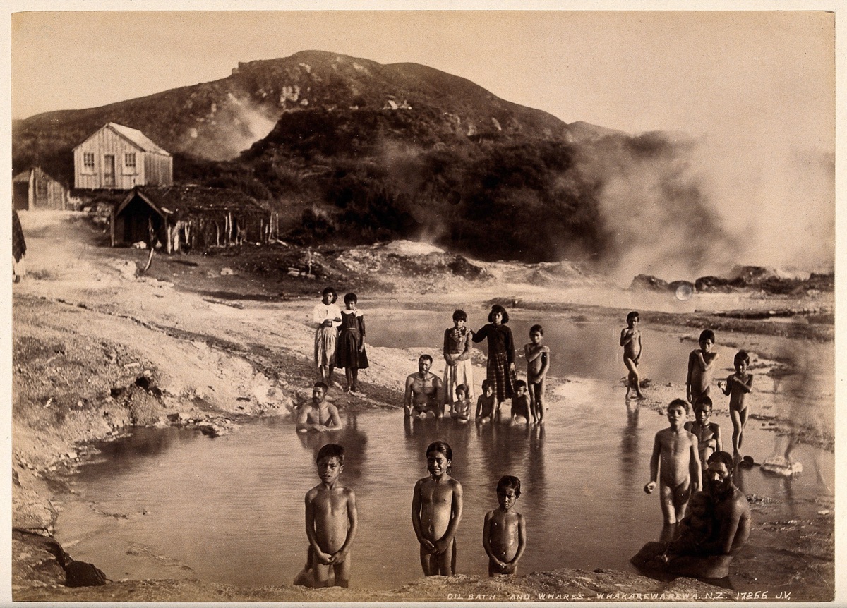 Whakarewrewa, New Zealand: a group of Maori people, thermal baths, and houses. Albumen print.